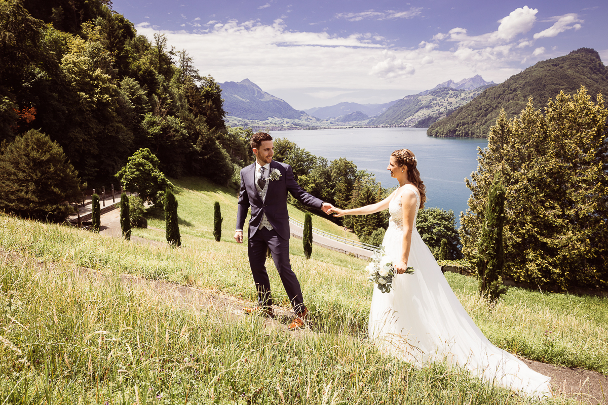 Brautpaar spaziert Hand in Hand mit Blick auf einen See und die umliegenden Berge, Hochzeit im Freien