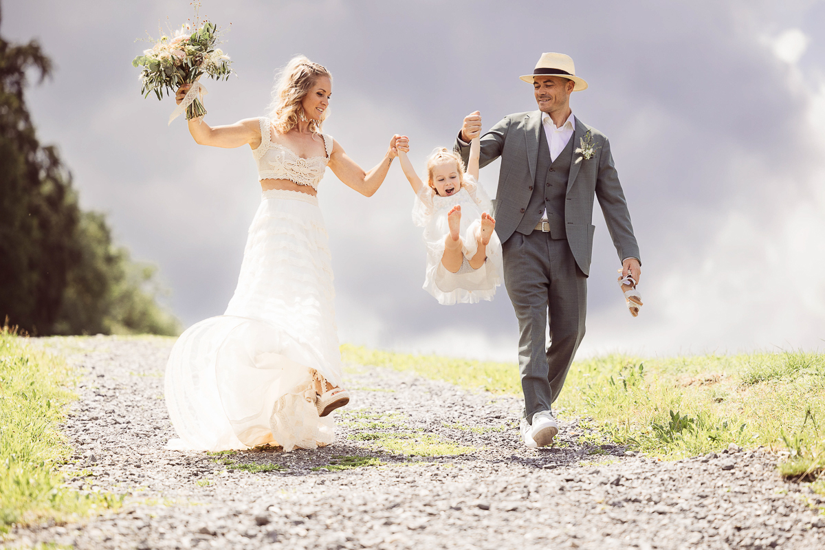 Brautpaar spaziert fröhlich mit ihrer Tochter an der Hand, Hochzeit im Freien