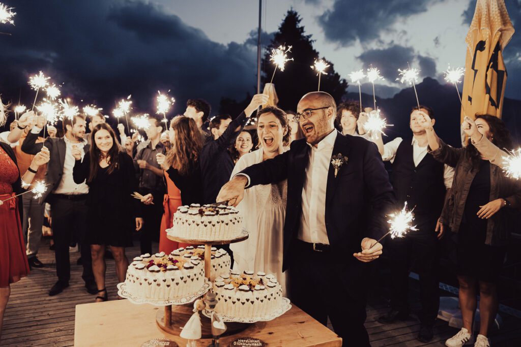 Brautpaar beim Anschnitt der Hochzeitstorte, umgeben von Gästen mit Wunderkerzen, aufgenommen auf der Alp Flix von Hochzeitsfotograf Adrian Flütsch.