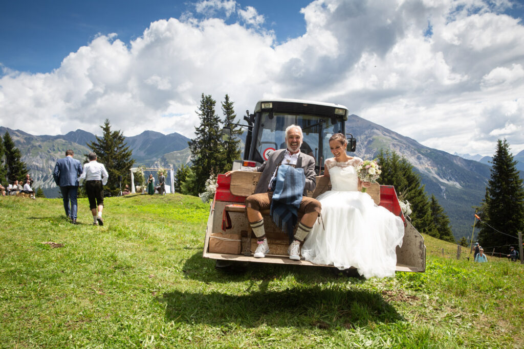 Brautvater begleitet seine Tochter auf einem Traktor zur Trauung während einer Alphochzeit in Lenzerheide, fotografiert von Hochzeitsfotograf Adrian Flütsch.