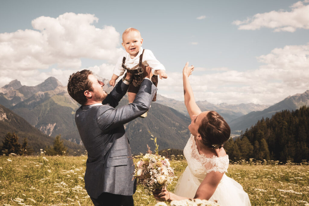 Hochzeit in Lenzerheide: Glückliches Brautpaar mit Sohn vor der Bergkulisse, eingefangen von Hochzeitsfotograf Adrian Flütsch.