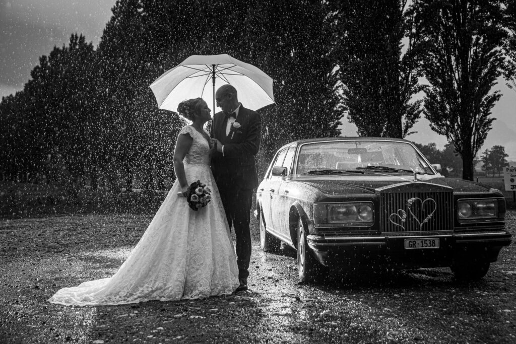 Schwarz-Weiß-Aufnahme des Hochzeitspaares im Regen mit Regenschirm und Rolls-Royce – Adrian Flütsch