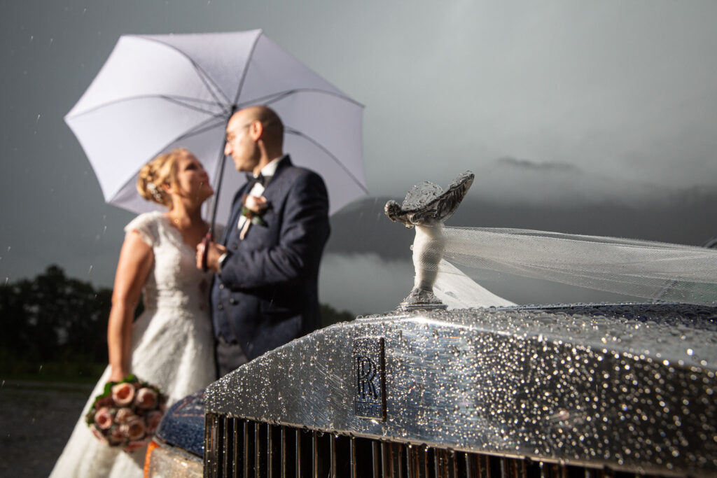 Detailaufnahme von Rolls-Royce mit Hochzeitspaar und Regenschirm im Hintergrund – Adrian Flütsch